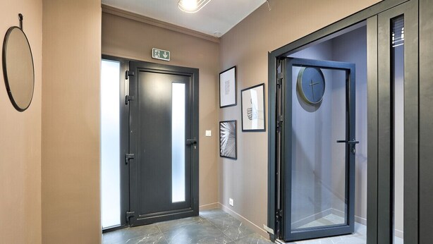 A hallway with a dark door, a glass door with a clock, and framed artwork on the wall.