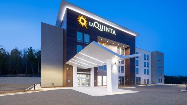 A La Quinta hotel with a modern design, featuring a prominent sun logo, a covered entrance, and a spacious parking area. - La Quinta by Wyndham Chelsea Birmingham (Chelsea)