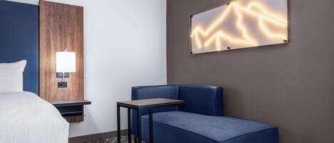 A modern hotel room with a bed, a desk, a blue sofa, a wall-mounted lamp, and a decorative light panel.