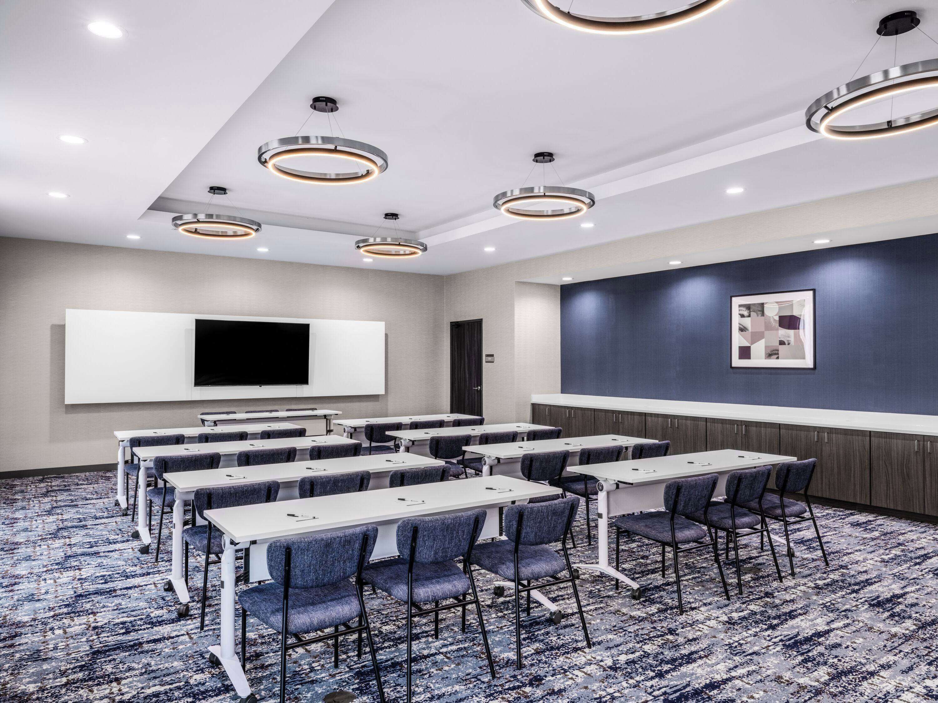 A conference room with a large flat-screen TV, multiple tables with chairs, and modern circular ceiling lights.