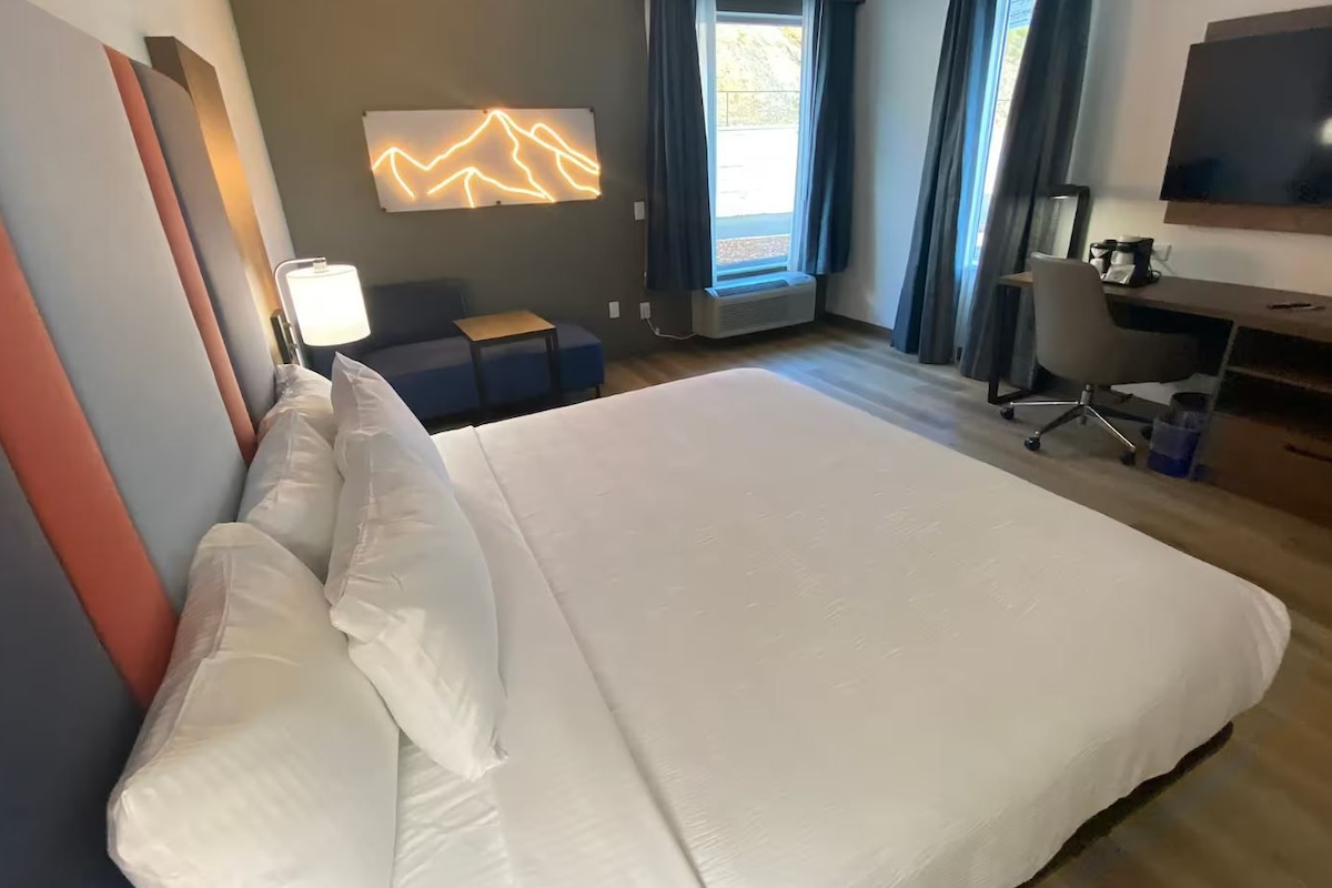 A hotel room with a large bed, a desk with a chair, a flat-screen TV, and a window with curtains.