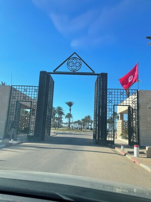 A gated entrance with a red flag, palm trees, and a clear blue sky.
