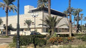A modern building with a flat roof, surrounded by palm trees and a well-maintained garden with orange flowers.