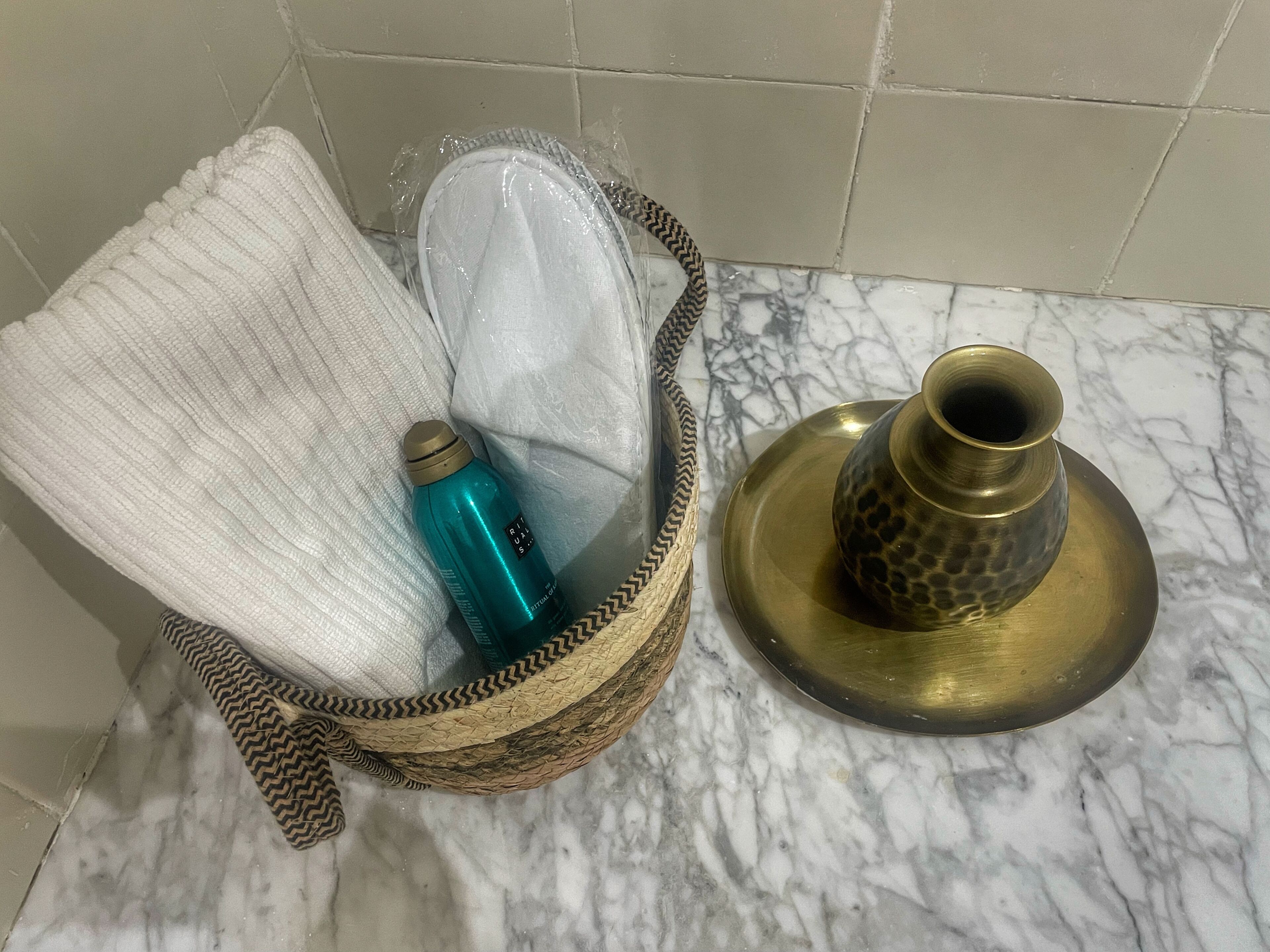 Un baño con piso de mármol, una cesta tejida que contiene una toalla y una botella de agua, y un recipiente de latón sobre una bandeja.