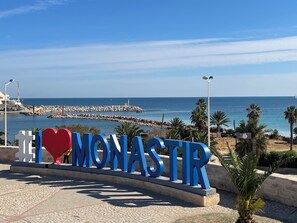A sign with 'I ♥ MONASTIR' in blue letters, a rocky breakwater, and a clear blue sky.