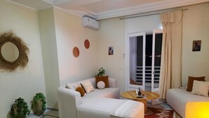 A modern living room with a sofa, a coffee table, and a bed. There is a balcony with curtains and a view of the city at night.