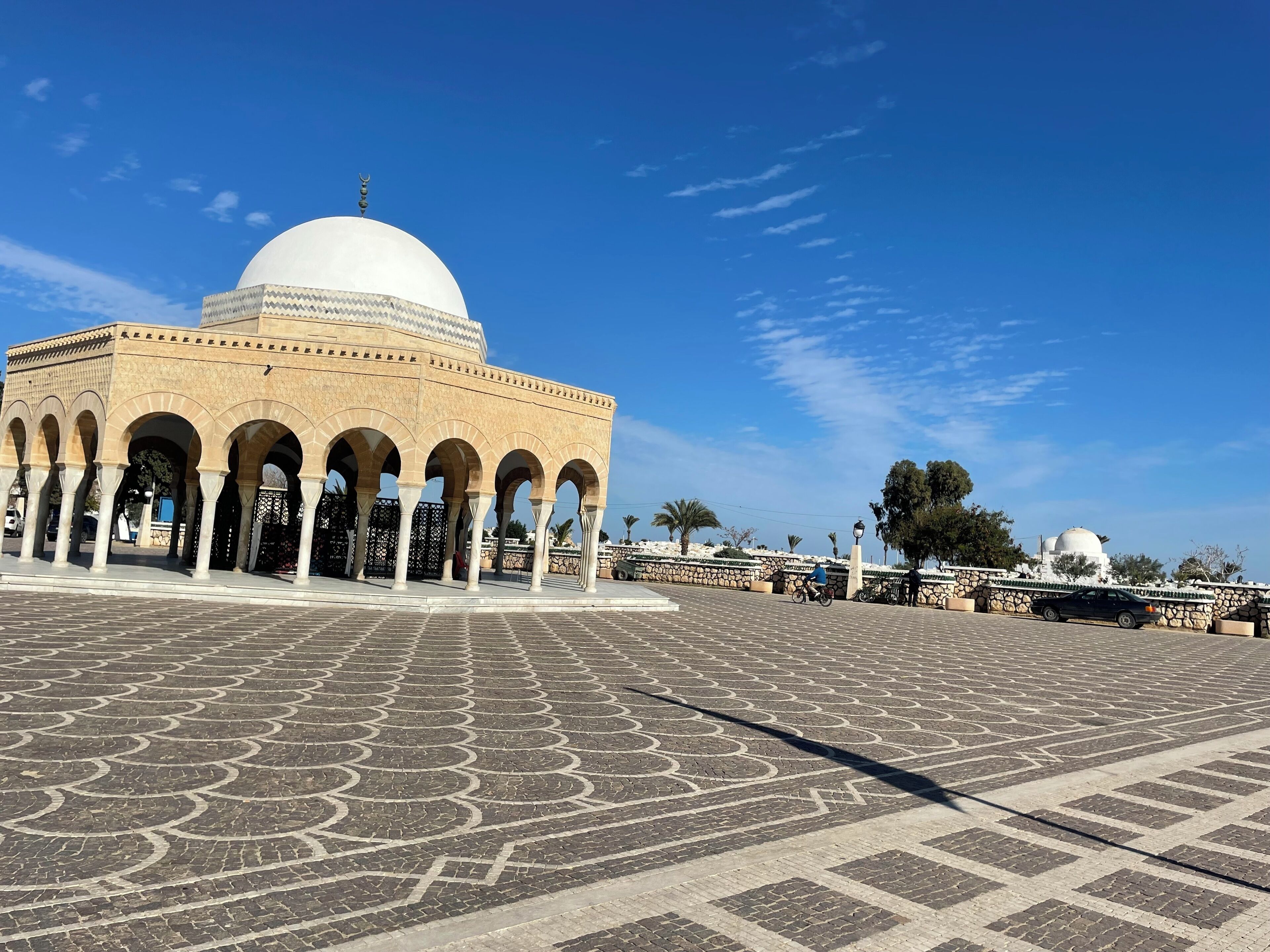 A mosque with a large dome and multiple arches, a paved courtyard, and a stone wall in the background.