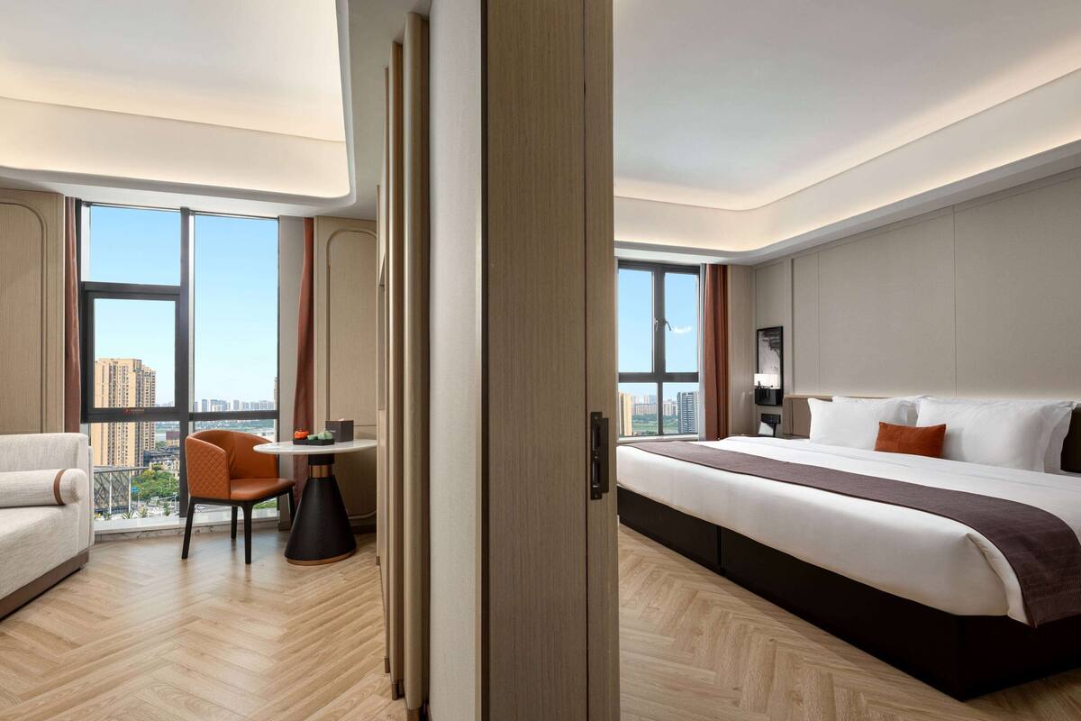 A modern hotel room with a large bed, a sofa, a dining table, and a view of the city.