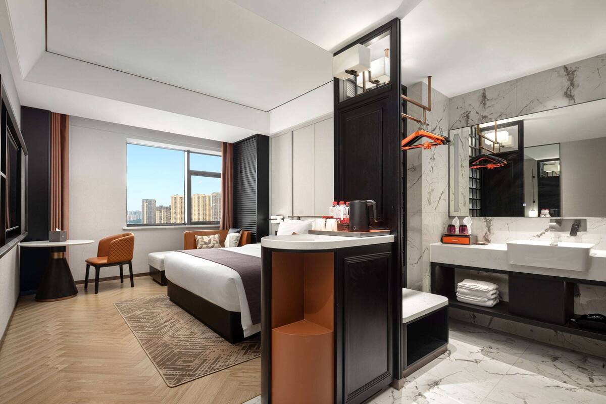 A modern hotel room with a bed, a desk, a chair, a bathroom with a mirror, and a view of the city.