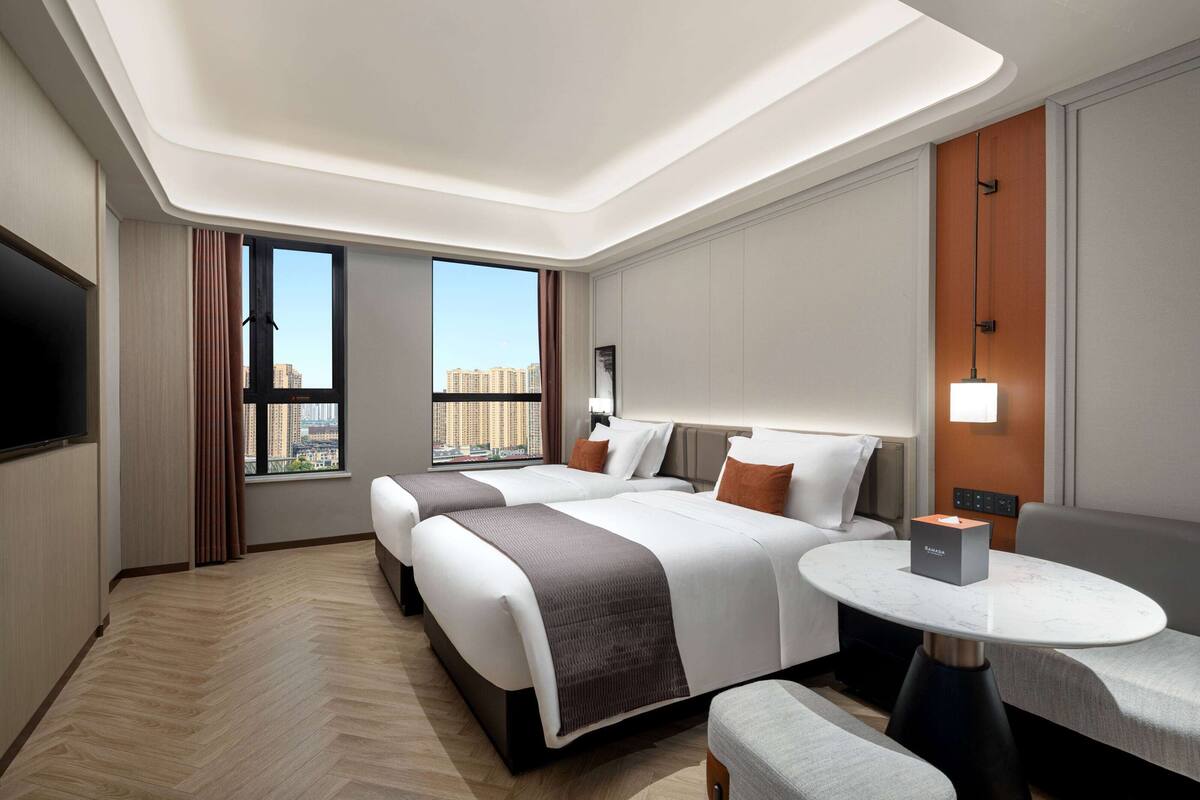 A hotel room with two beds, a round dining table, a TV, and a large window with a city view.