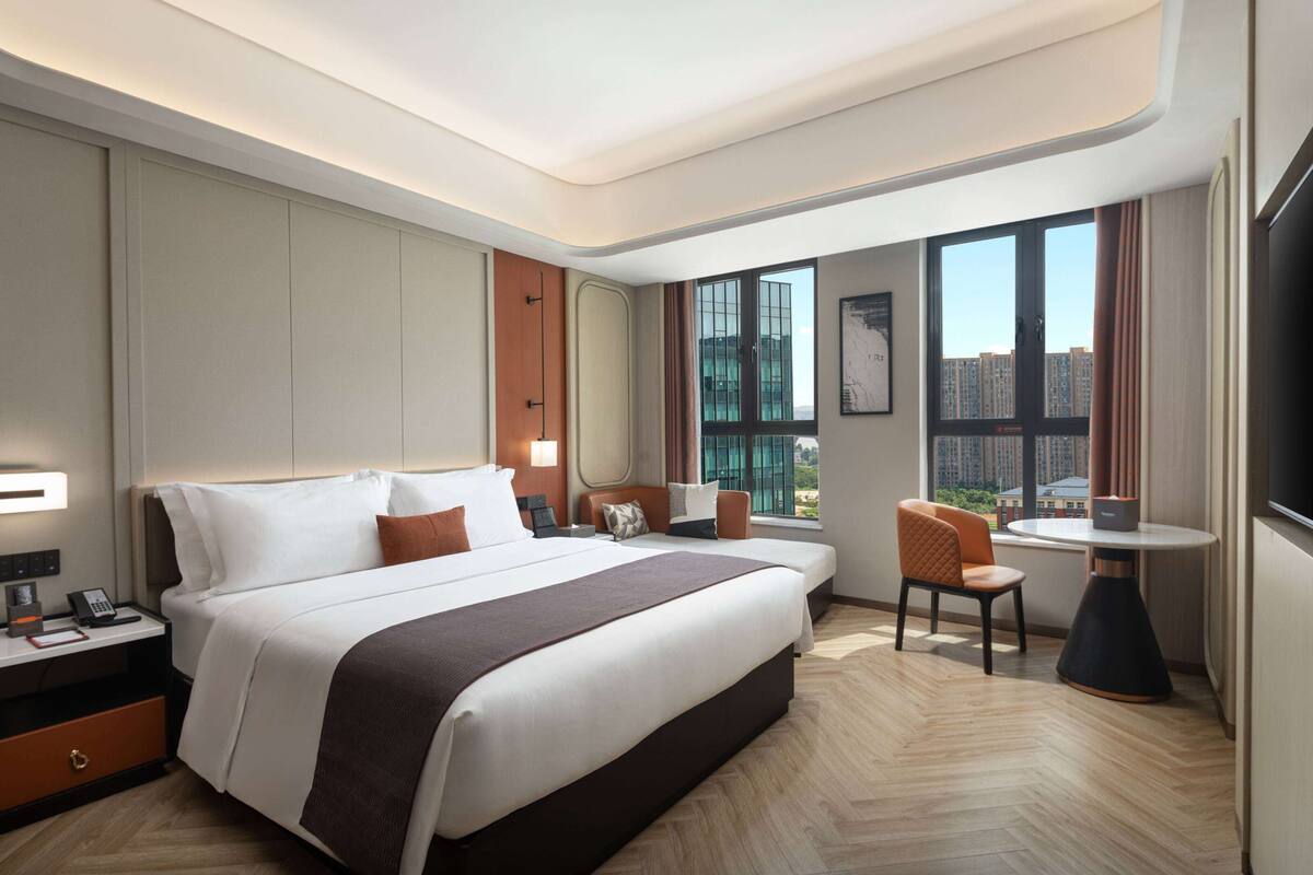 A modern hotel room with a large bed, a desk with a chair, a small table, and a large window offering a city view.