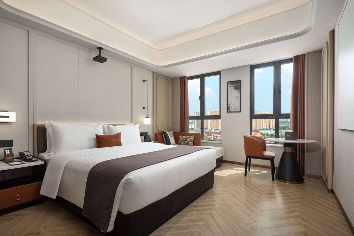 A modern hotel room with a large bed, a desk with a phone, a chair, and a view of the city through large windows.