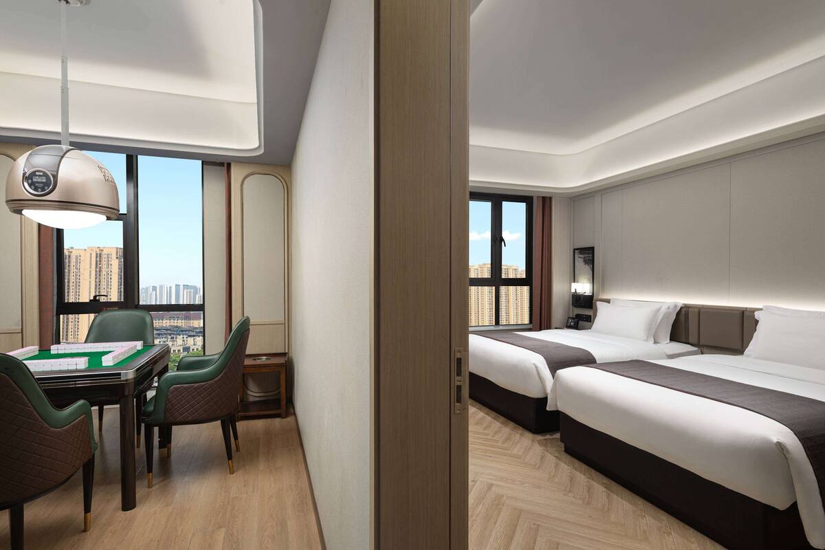 A hotel room with a desk, chairs, a bed, and a view of the city.