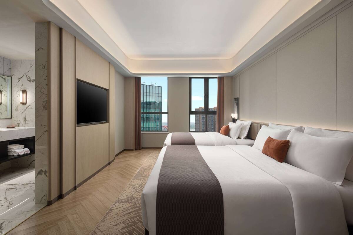 A hotel room with a large bed, a flat-screen TV, a marble bathroom vanity, and a city view through the windows.
