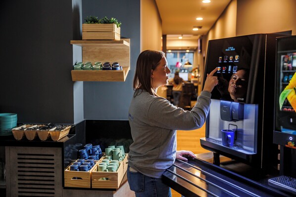 Uma pessoa interage com um quiosque digital em um ambiente moderno, com prateleiras de madeira e uma máquina de café.