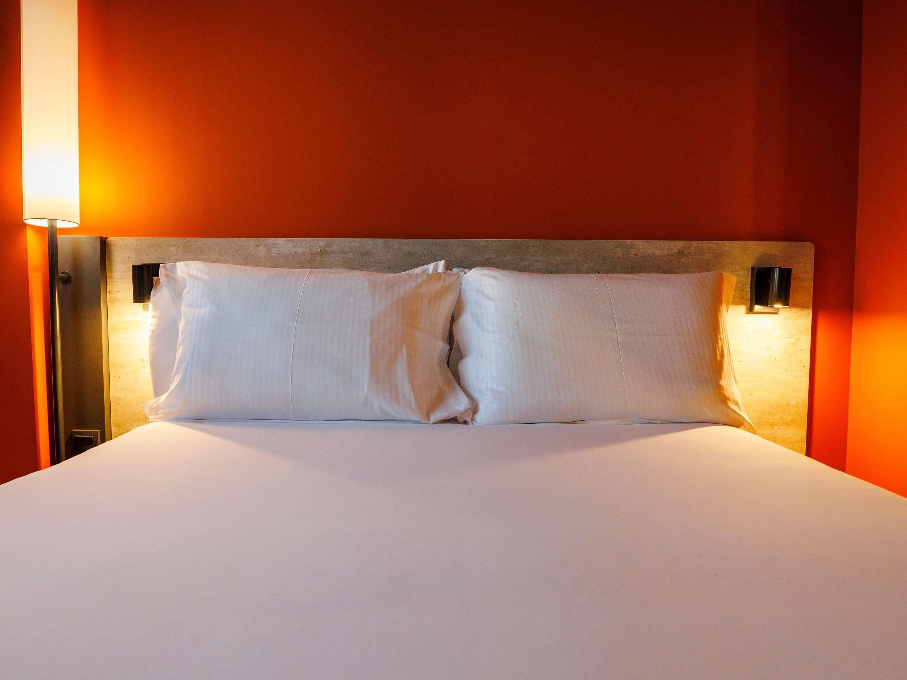 A hotel room with a bed, two pillows, a nightstand, and a wall-mounted lamp.