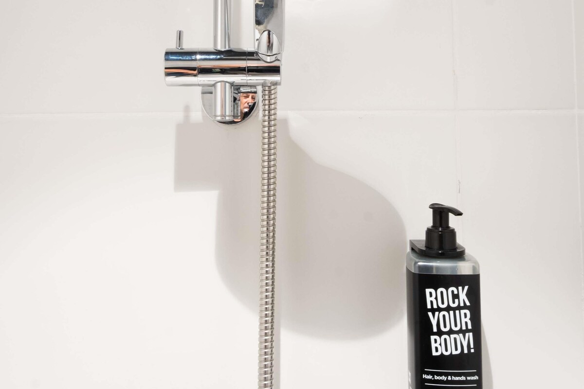 A shower with a chrome faucet and a handheld showerhead. A black bottle with the text 'ROCK YOUR BODY!' is placed beside it.