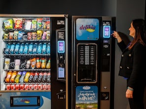 Un distributeur automatique proposant diverses collations et boissons, une machine à café, et une femme interagissant avec ces appareils.