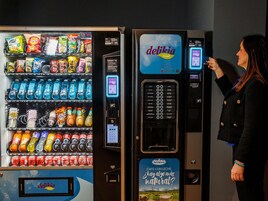 Máquina expendedora con diferentes aperitivos y bebidas, una máquina de café y una mujer usándola.