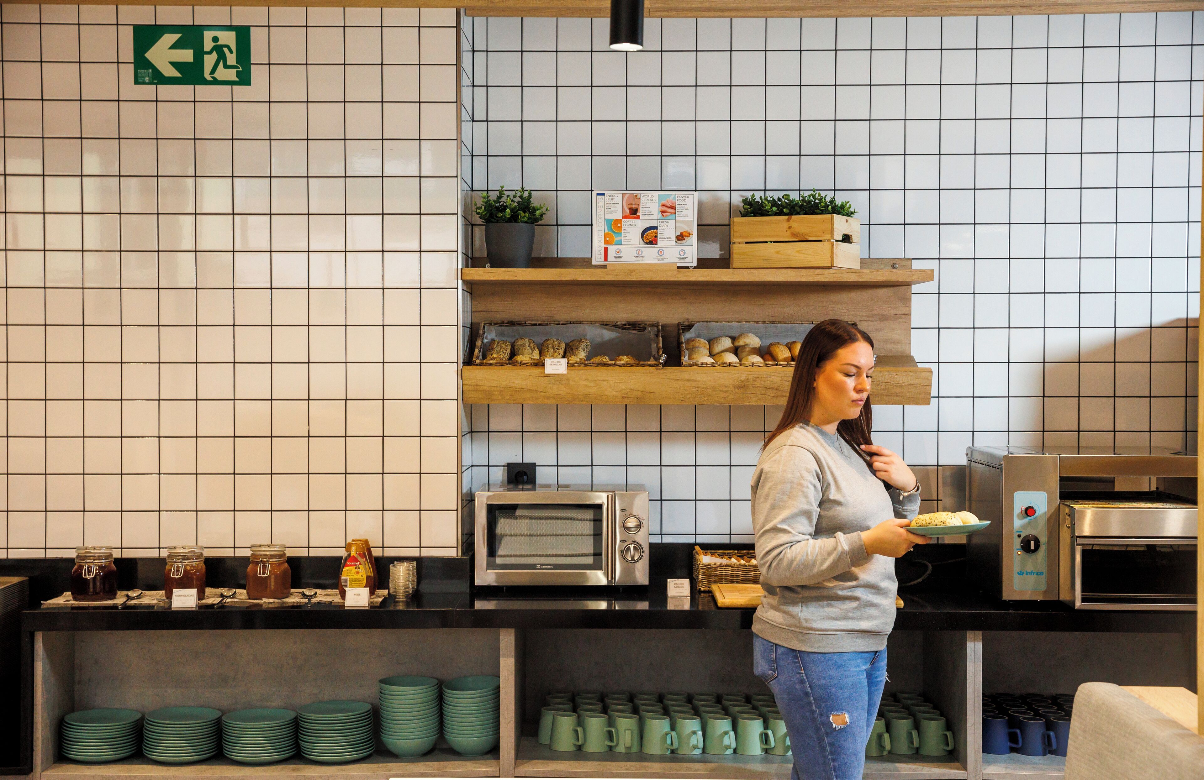 A kitchen area with a woman holding a plate of food, a microwave, a toaster, and shelves with jars and mugs.