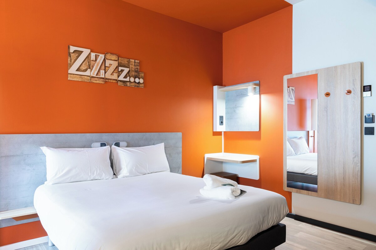 A modern hotel room with a large bed, a wooden headboard, a wall-mounted mirror, and a bright orange accent wall.