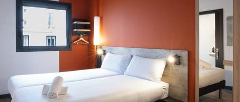 Habitación de hotel con una cama grande, una mesita de noche, una lámpara de pared, una ventana con vistas a edificios y un espejo.