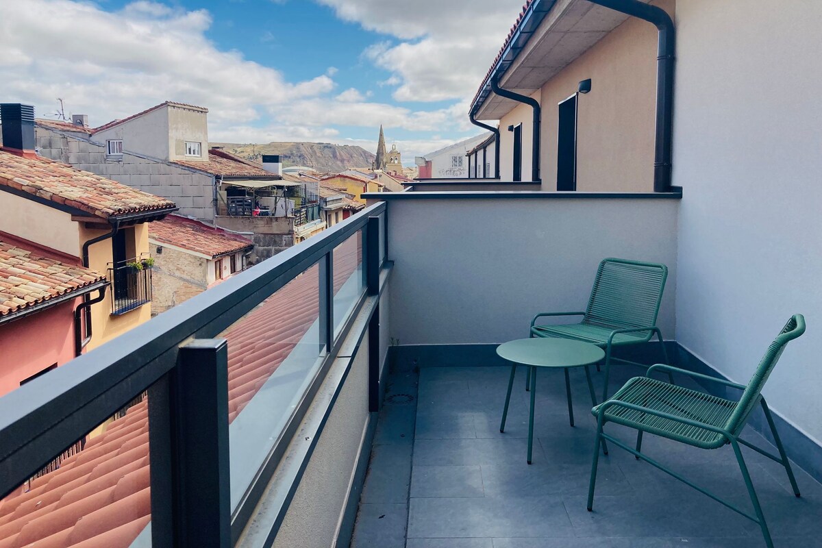 A balcony with two green chairs and a small table, overlooking a cityscape with traditional buildings and a church spire.