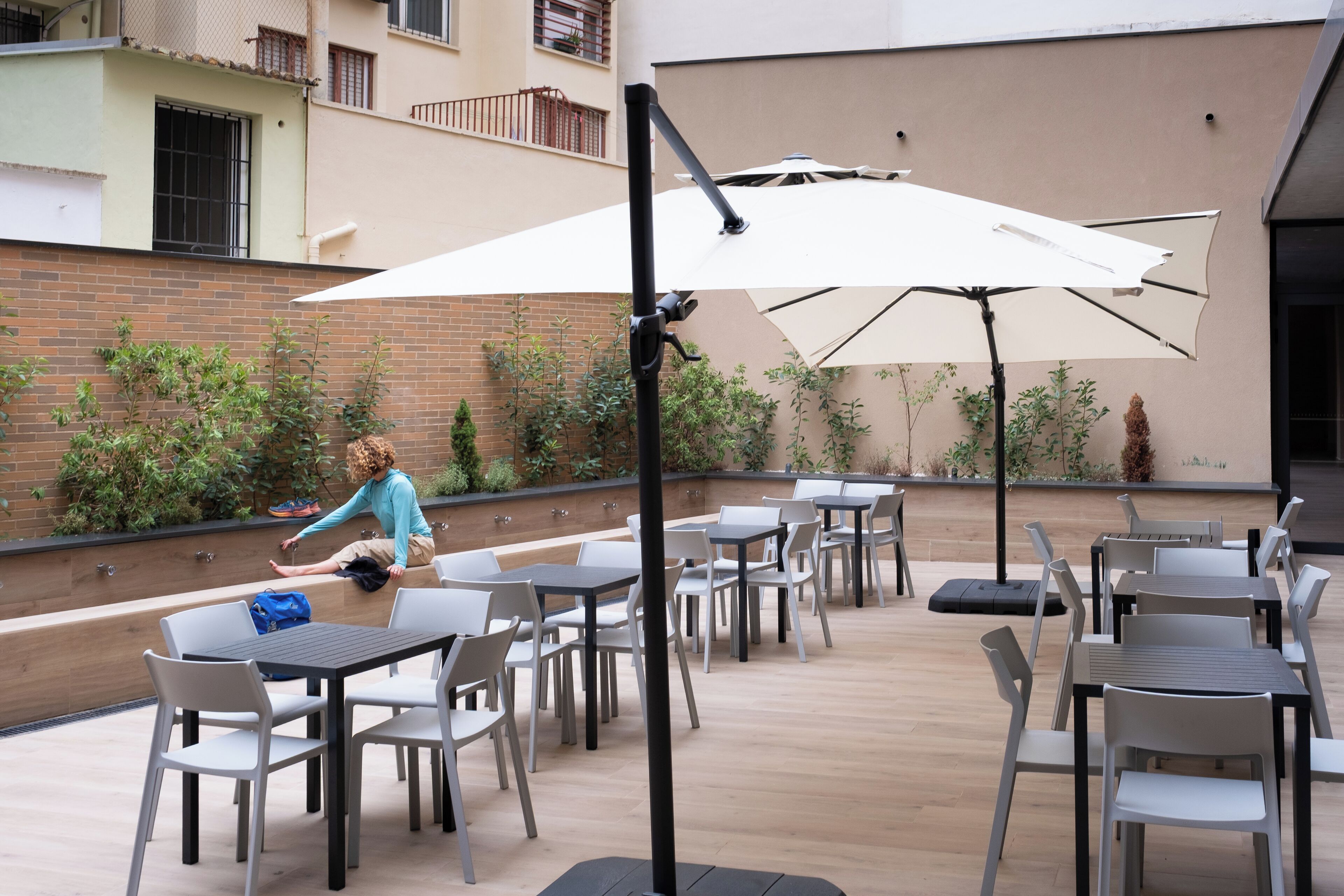 An outdoor patio with tables and chairs, a person sitting on a bench, and a large umbrella providing shade.