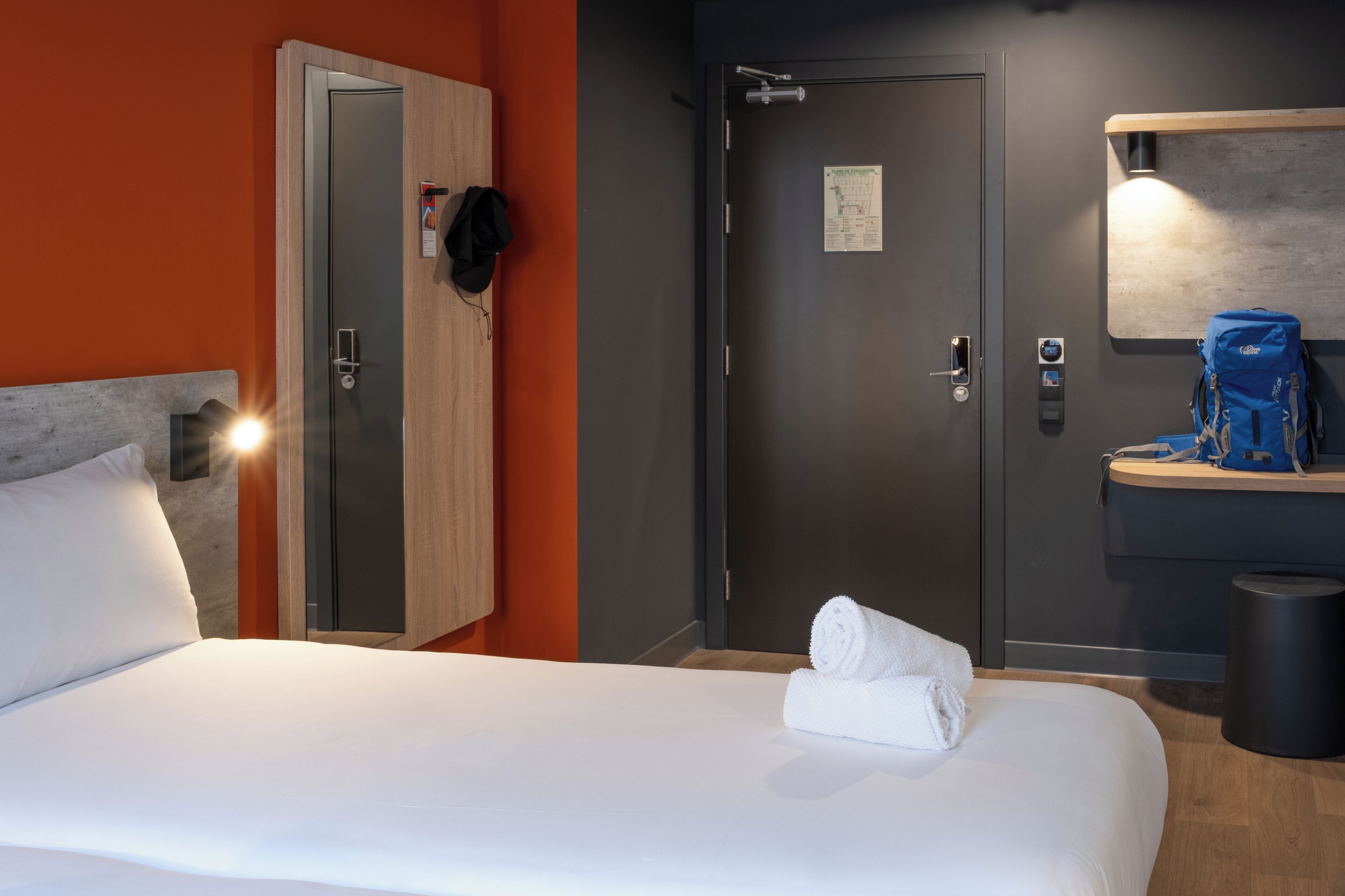 Une chambre d’hôtel avec un lit, une armoire, une porte, un miroir, une serviette, une poubelle et une petite table sur laquelle se trouve un sac.