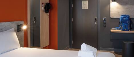 A hotel room with a bed, a wardrobe, a door, a mirror, a towel, a trash bin, and a small table with a bag.