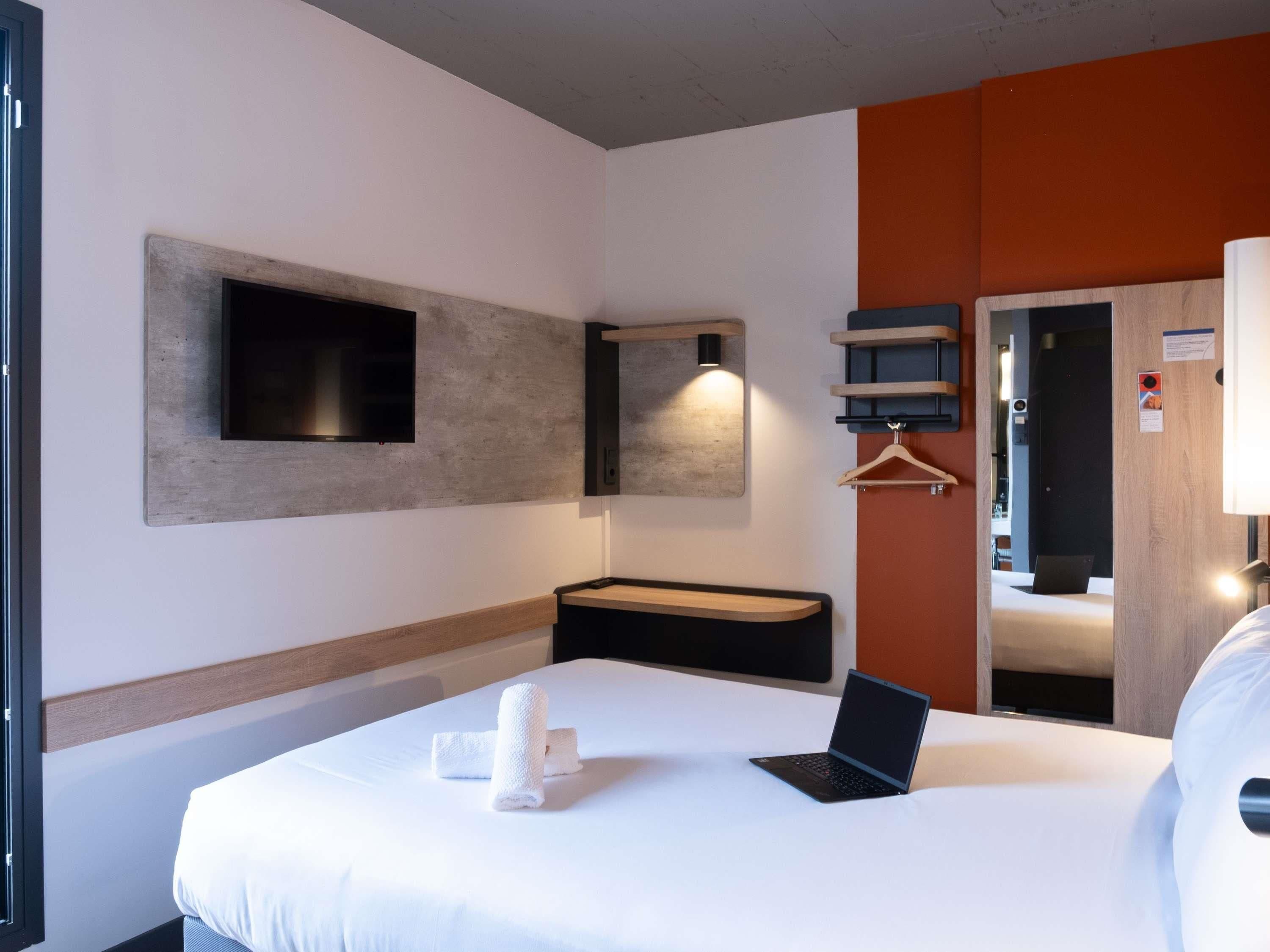 A hotel room with a bed, a laptop, a TV, a coat rack, and a mirror.