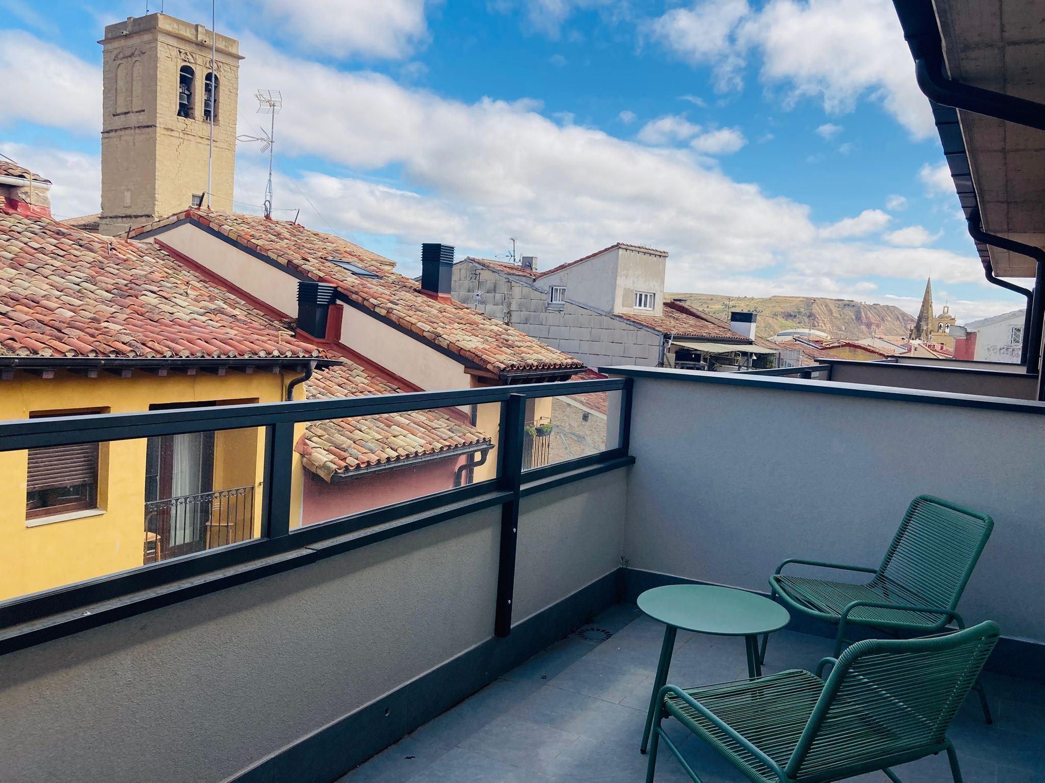 Un balcón con vistas a edificios de tejas rojizas, una pequeña mesa y dos sillas verdes.