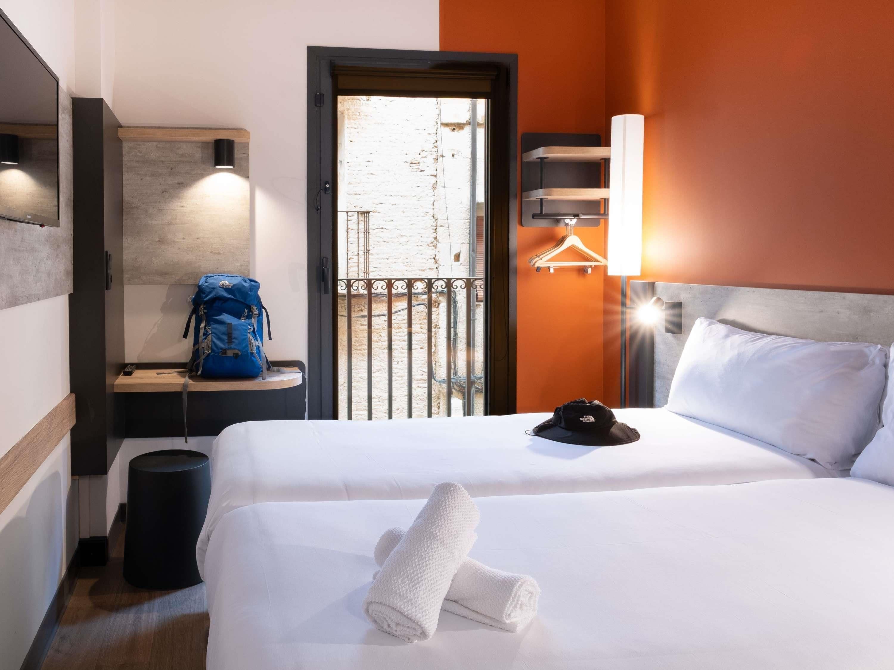 A hotel room with a bed, a small table with a backpack, a trash bin, a wall-mounted light, and a view of the outside balcony.