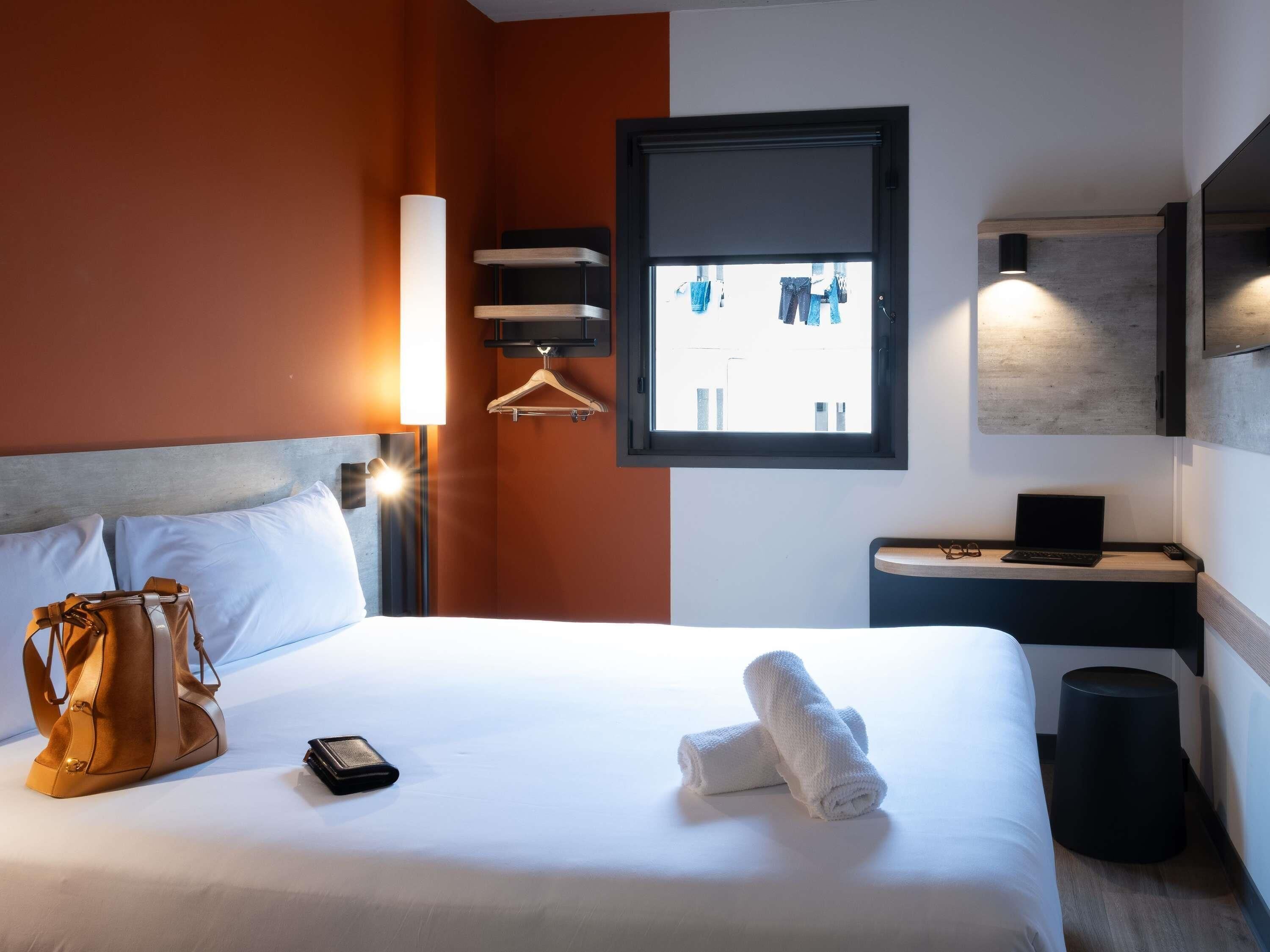 Ett hotellrum med en säng, en klädstång, ett skrivbord med en bärbar dator och ett fönster med utsikt.