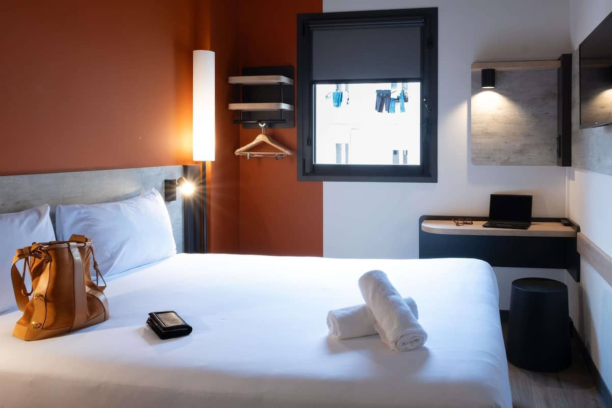 A hotel room with a bed, a hanging coat rack, a desk with a laptop, and a window with a view of buildings.