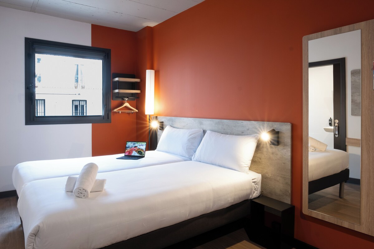 A hotel room with a large bed, a bedside table, a lamp, a window, and a mirror.