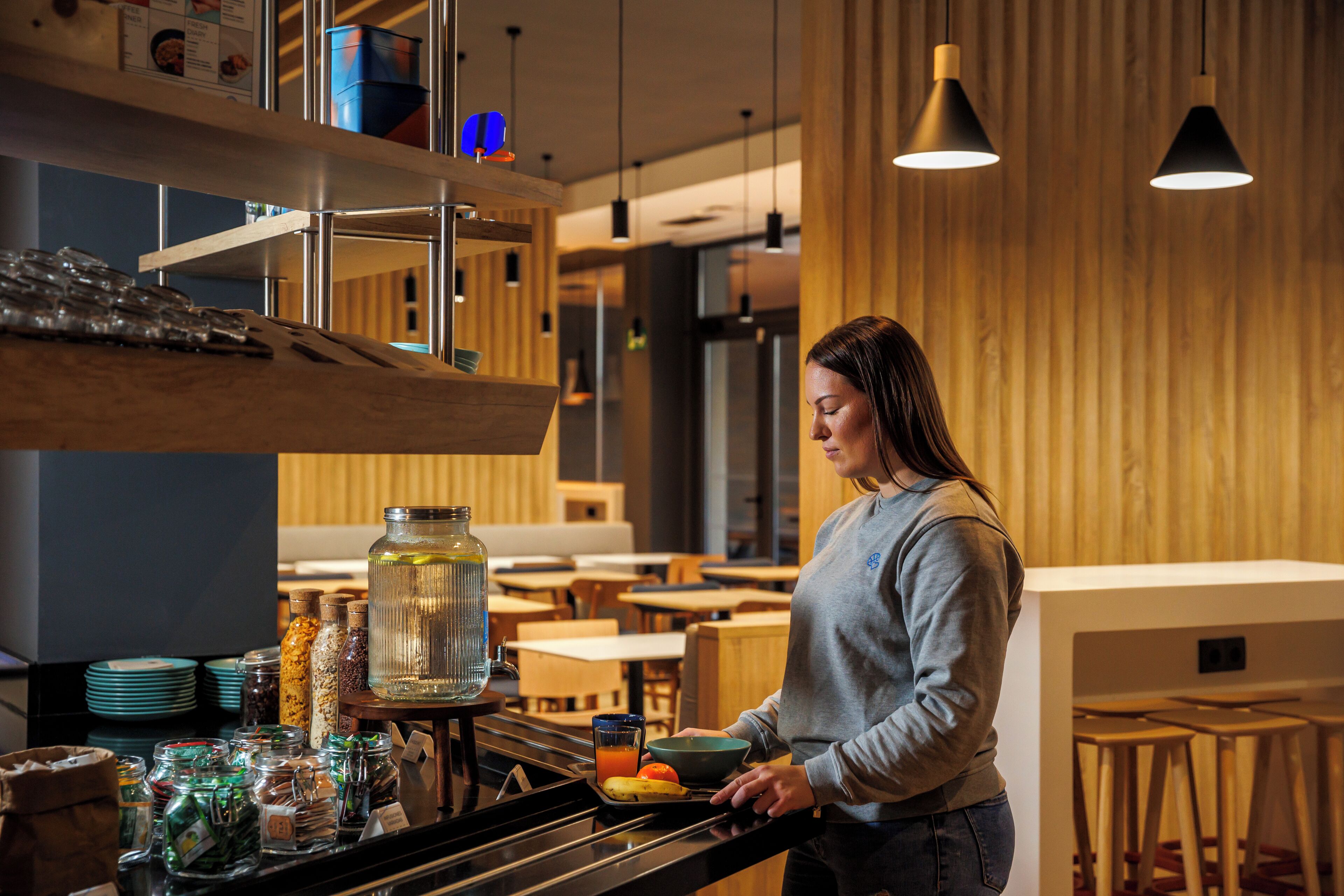 Una mujer preparando comida en una barra con diferentes condimentos y un dispensador de bebidas.