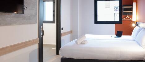 A hotel room with a bed, a TV mounted on the wall, a window, and a door leading to another room.