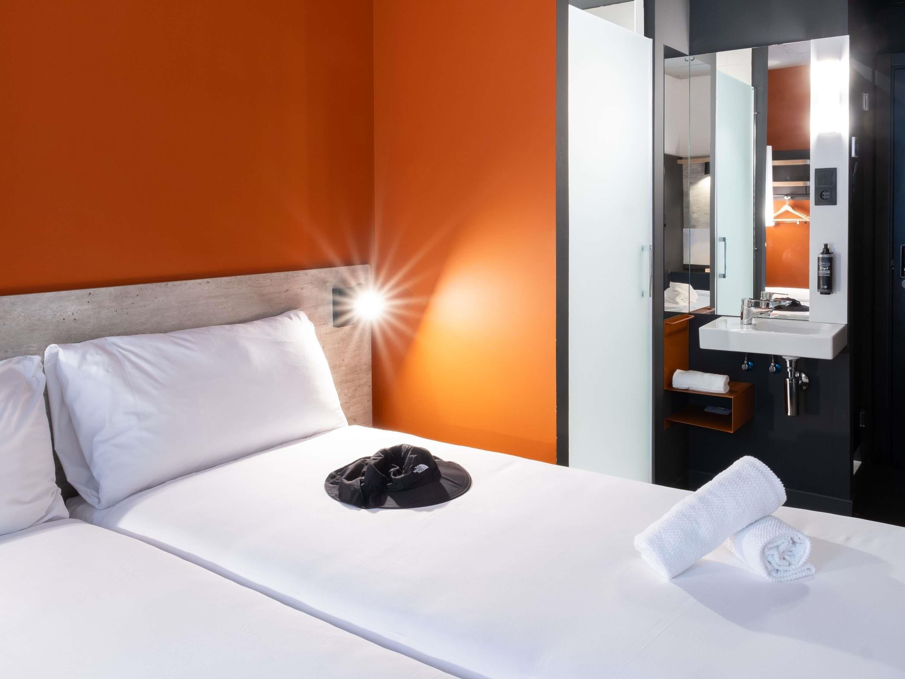 Une chambre d’hôtel avec un lit, un oreiller blanc, une casquette noire et une serviette enroulée.