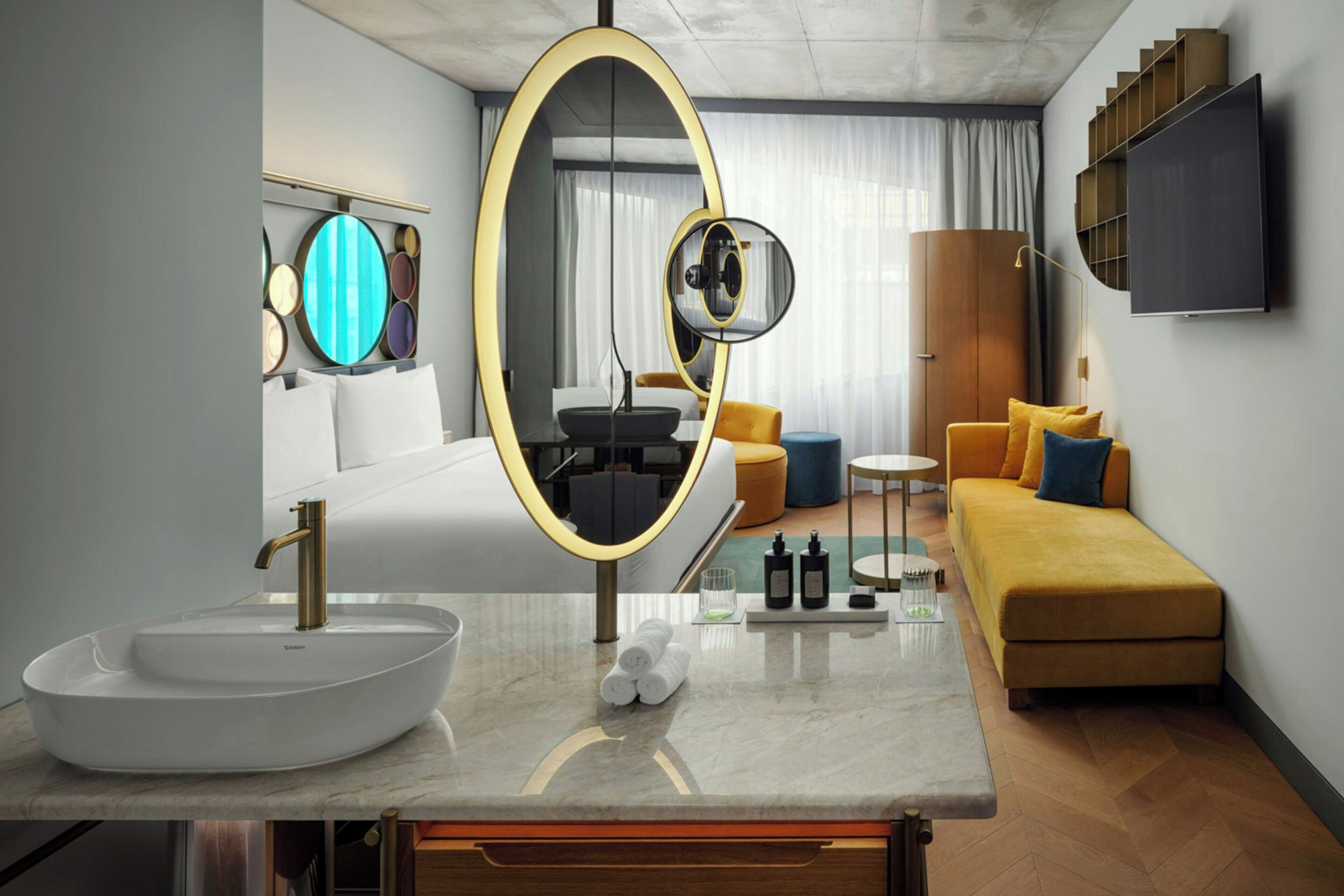 A modern hotel room with a large oval mirror, a white sink, a sofa, and a flat-screen TV mounted on the wall.