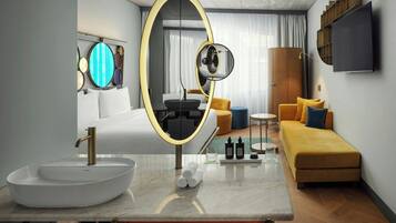 A modern hotel room with a large oval mirror, a white sink, a sofa, and a flat-screen TV mounted on the wall.