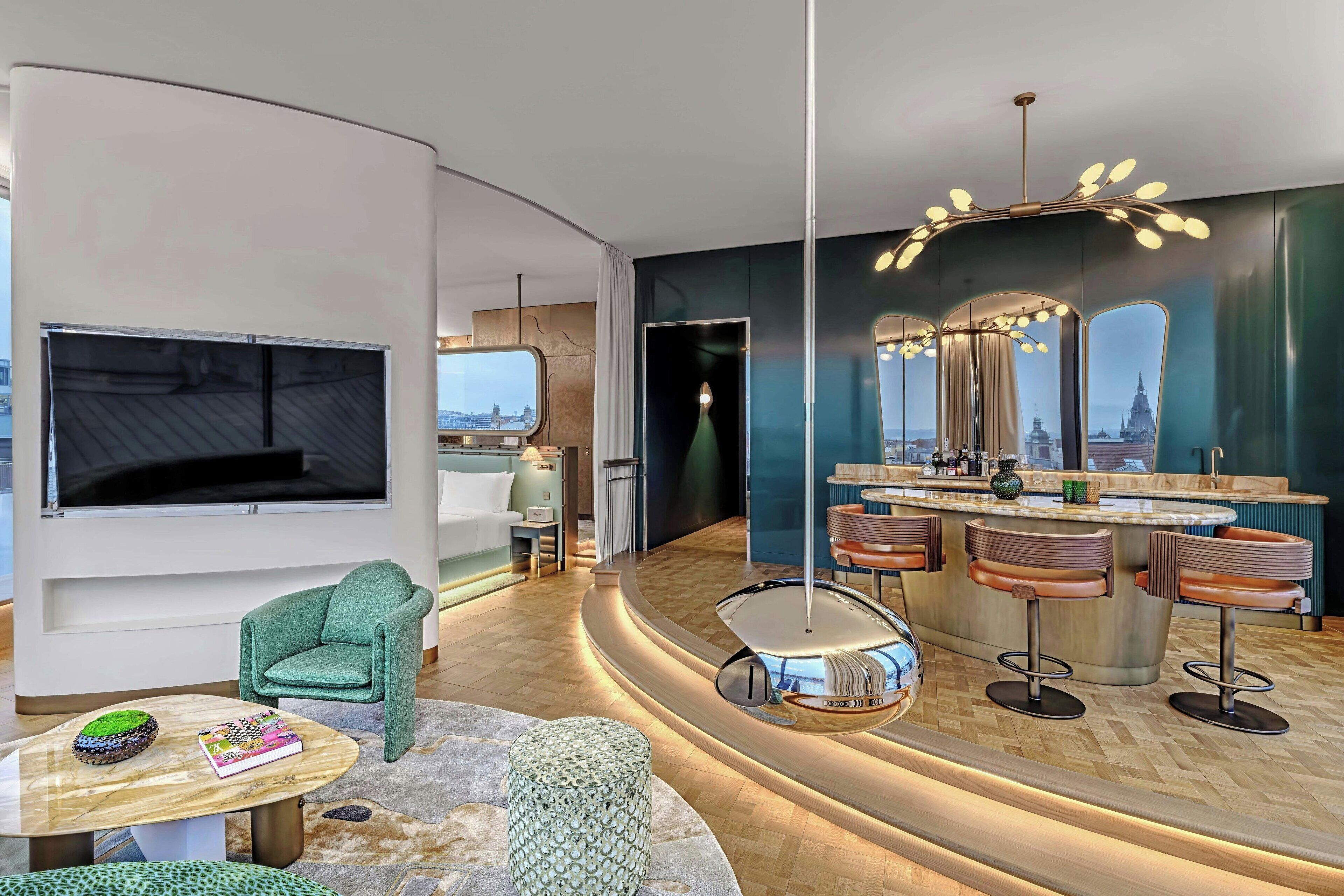 A modern hotel room with a curved layout, a large TV, a seating area with a green armchair, a small table with books, a unique cylindrical ottoman, and a bar area with stools and a mirror.