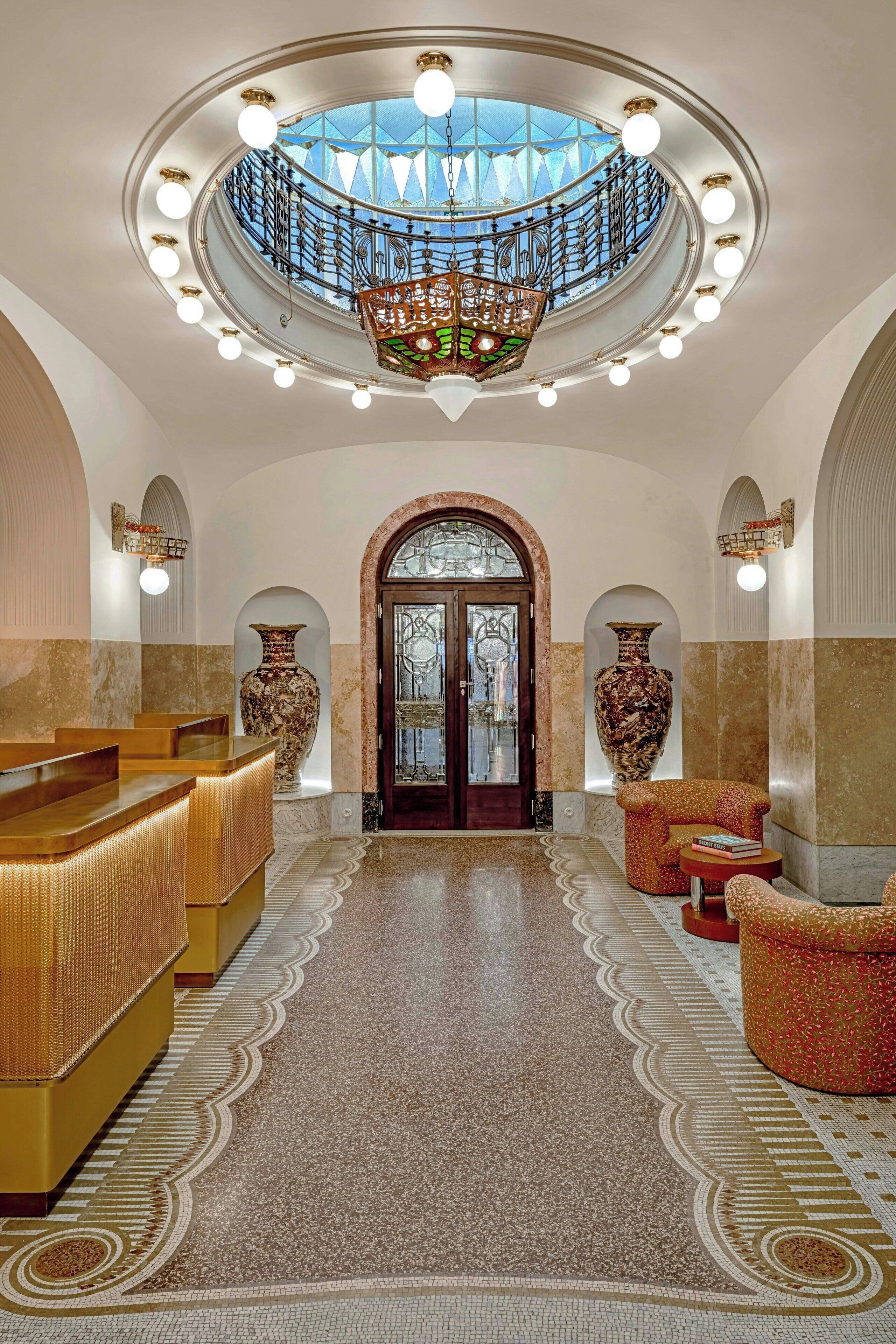Un vaste hall d’entrée avec un escalier central, de somptueux lustres et de grands vases.