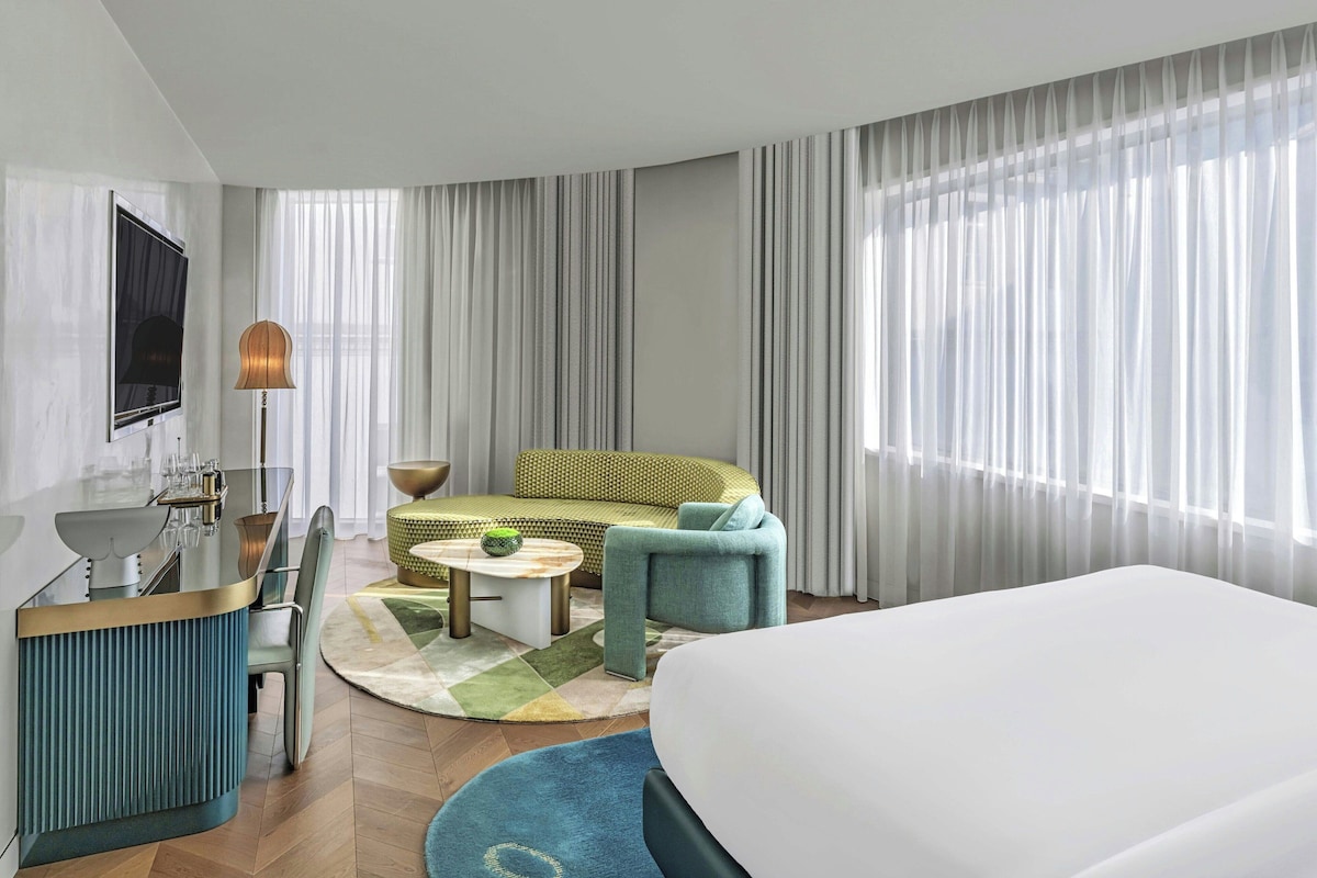 Habitación de hotel con cama, sofá, mesa de centro, escritorio con lámpara y televisor.