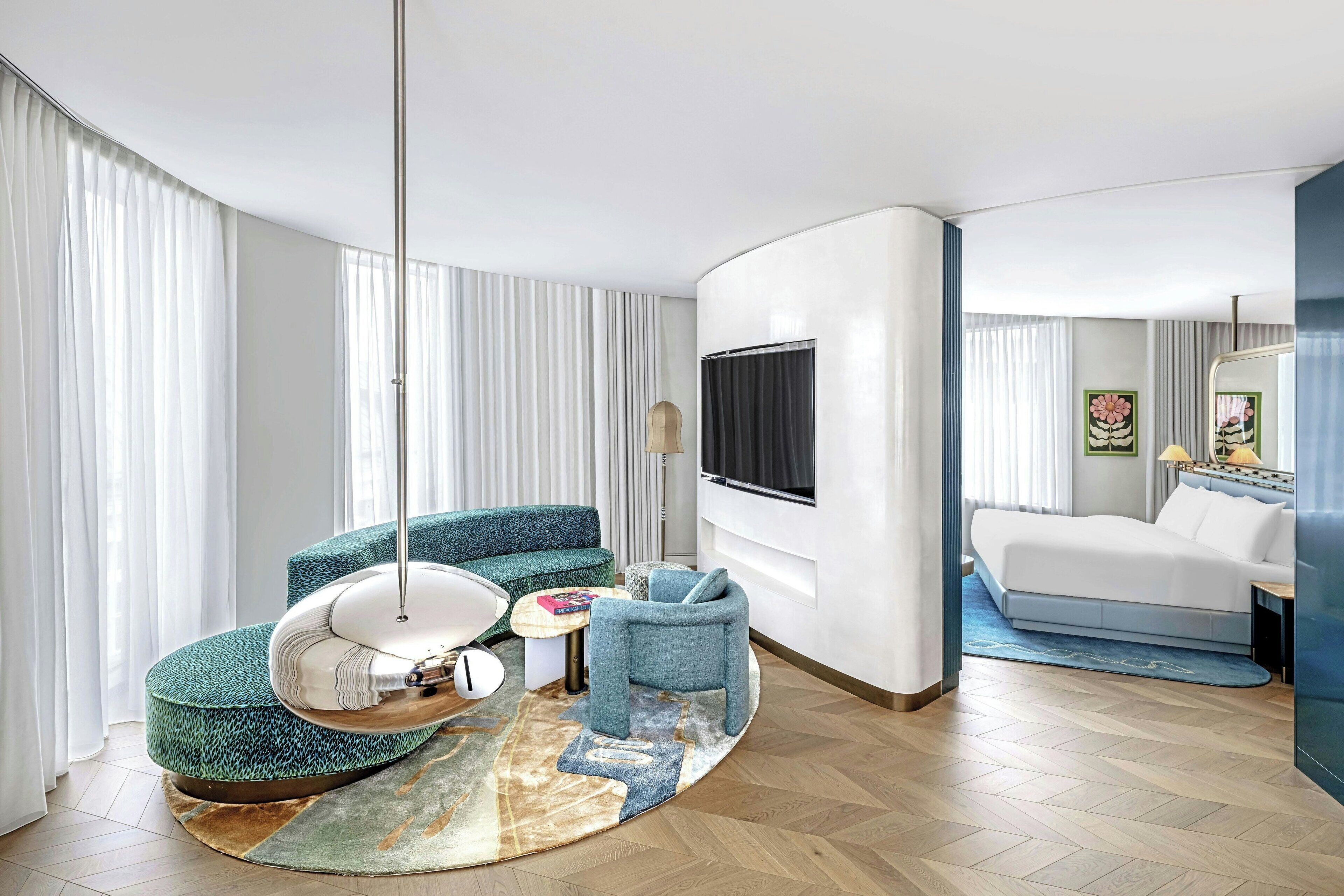 Ein modernes Hotelzimmer mit einer gebogenen Couch, einem runden Couchtisch, einem Hängekorbsessel, einem Bett und einem großen Fernseher.