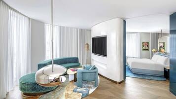 Ein modernes Hotelzimmer mit einer gebogenen Couch, einem runden Couchtisch, einem Hängekorbsessel, einem Bett und einem großen Fernseher.