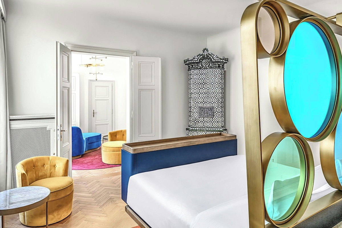 Una habitación de hotel con una cama, un espejo redondo, un sillón tapizado azul, una mesita y un pilar con diseño.