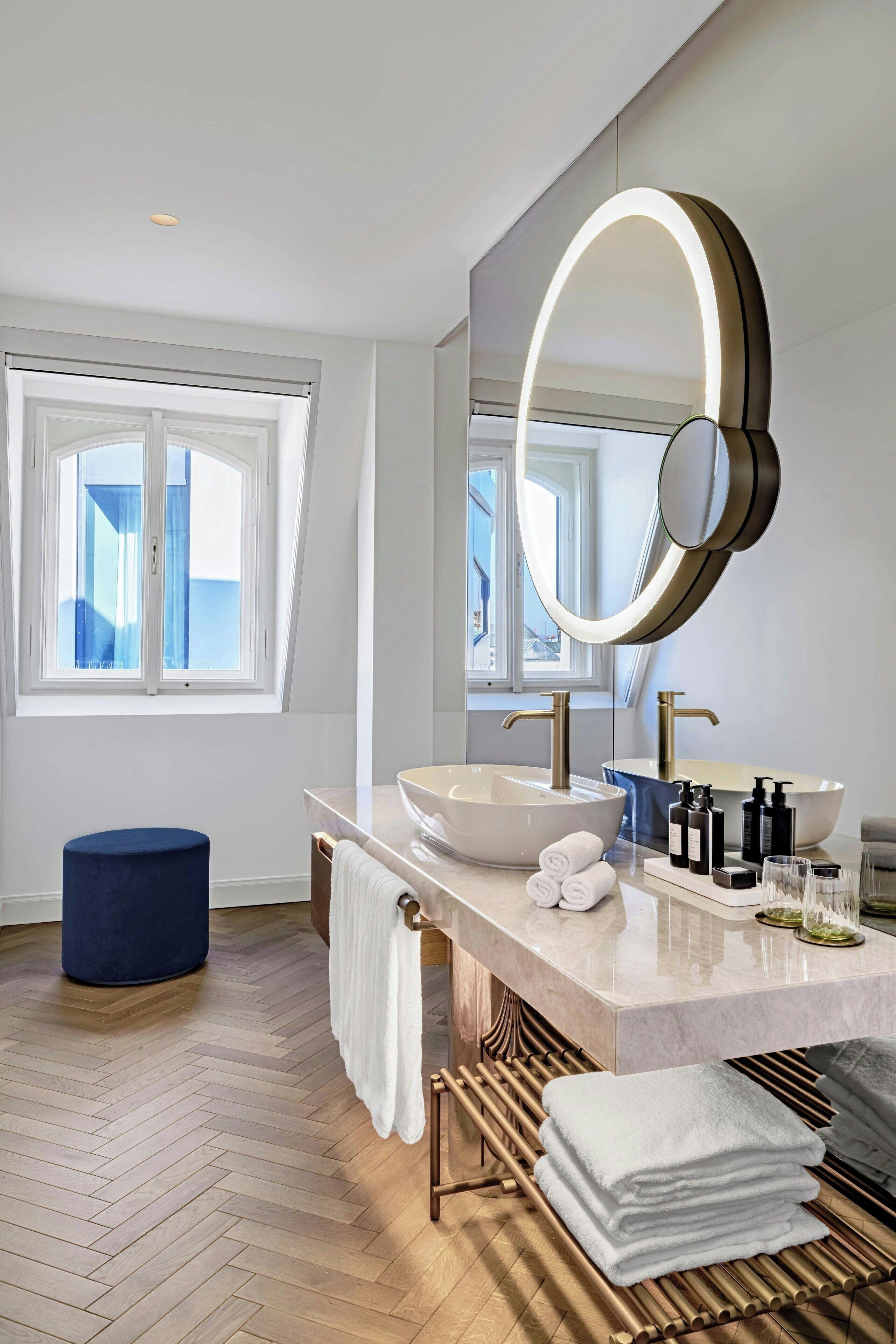 A modern bathroom with a large mirror, a white sink, and a blue ottoman.