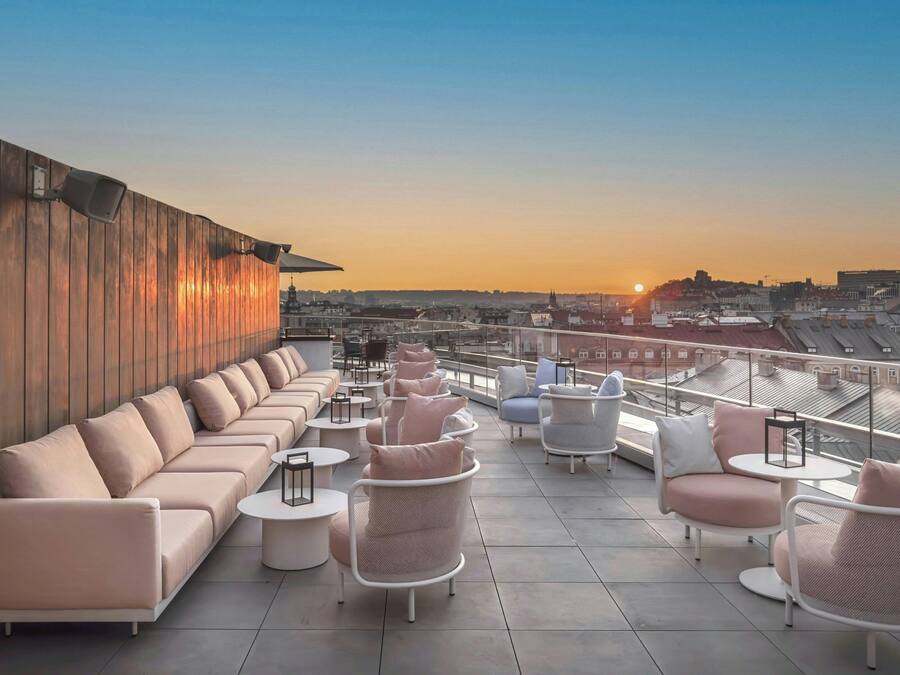 A rooftop terrace with a long sofa, round tables, and chairs, offering a view of the cityscape during sunset.