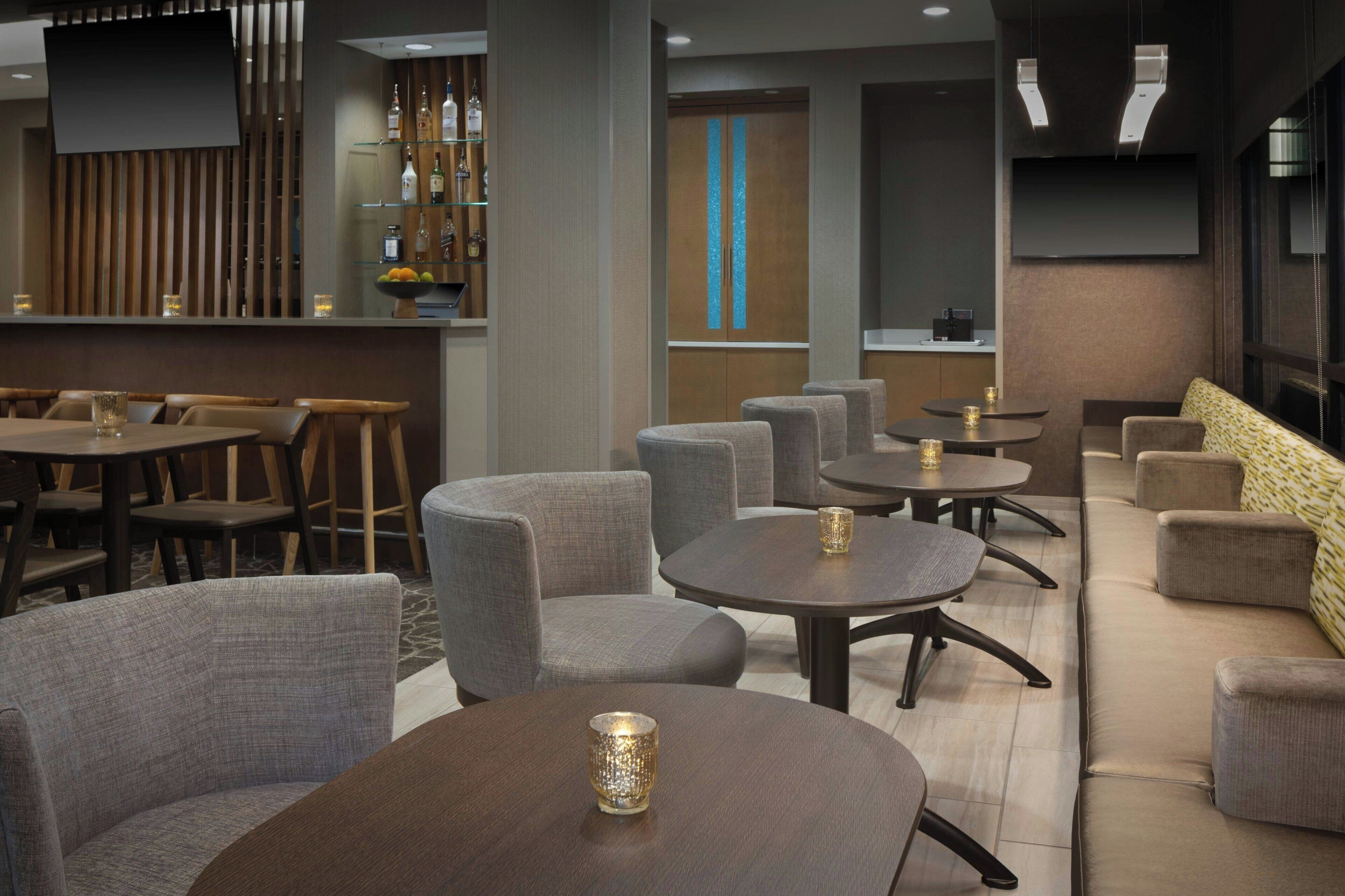 Ett modernt loungeområde med runda bord och bekväma sittplatser, en bar med en träpanel i bakgrunden och en stor TV-skärm.